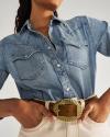 Person wearing a light denim button-up shirt with two chest pockets and a large rectangular gold belt buckle over light-colored pants.