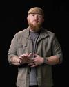 A bearded man wearing a tan cap, gray jacket, and smart watch stands against a black background, gesturing with his hands.
