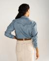 Woman standing with her back to the camera, wearing a light denim shirt, beige skirt with back pockets, and a brown belt.