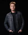 A man with short, light brown hair wears a dark jacket over a black shirt, standing against a black background with his hands in his jacket pockets.
