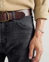 A person wearing a tan shirt, brown leather belt, dark jeans, and a silver chain bracelet, with their hand resting by their side.