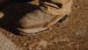 A close-up of a brown leather work boot and tan pants, both covered in dried mud, standing on a muddy, textured ground.