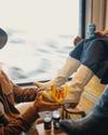 A person in a cowboy hat polishes white boots worn by another person sitting and holding a drink. Another person sitting nearby wears black boots.