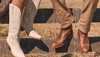 Close-up of red cowboy boots with intricate stitching, worn by someone standing on a textured surface. Another person wearing black and red cowboy boots is partly visible beside them.