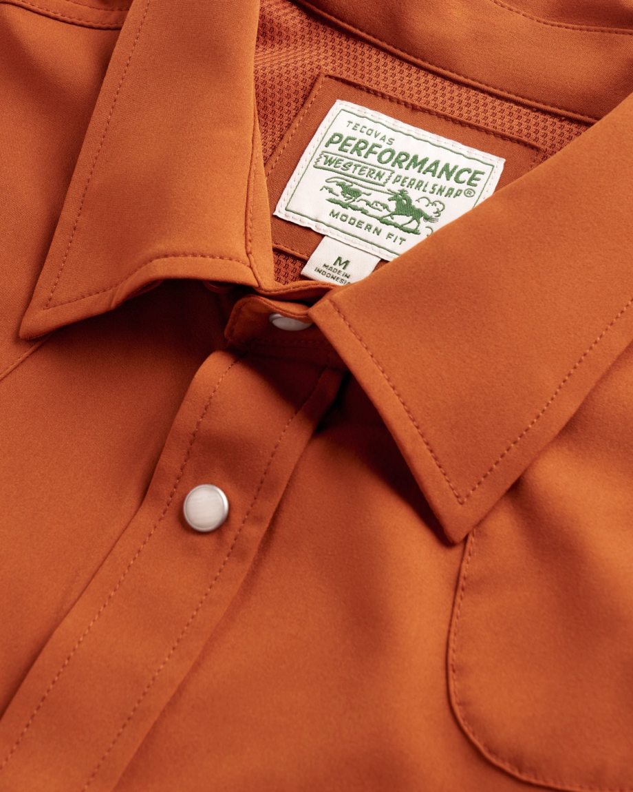 A close-up of a rust-colored Tecovas performance western pearl snap shirt, showing the collar, front snap button, and internal size medium label.