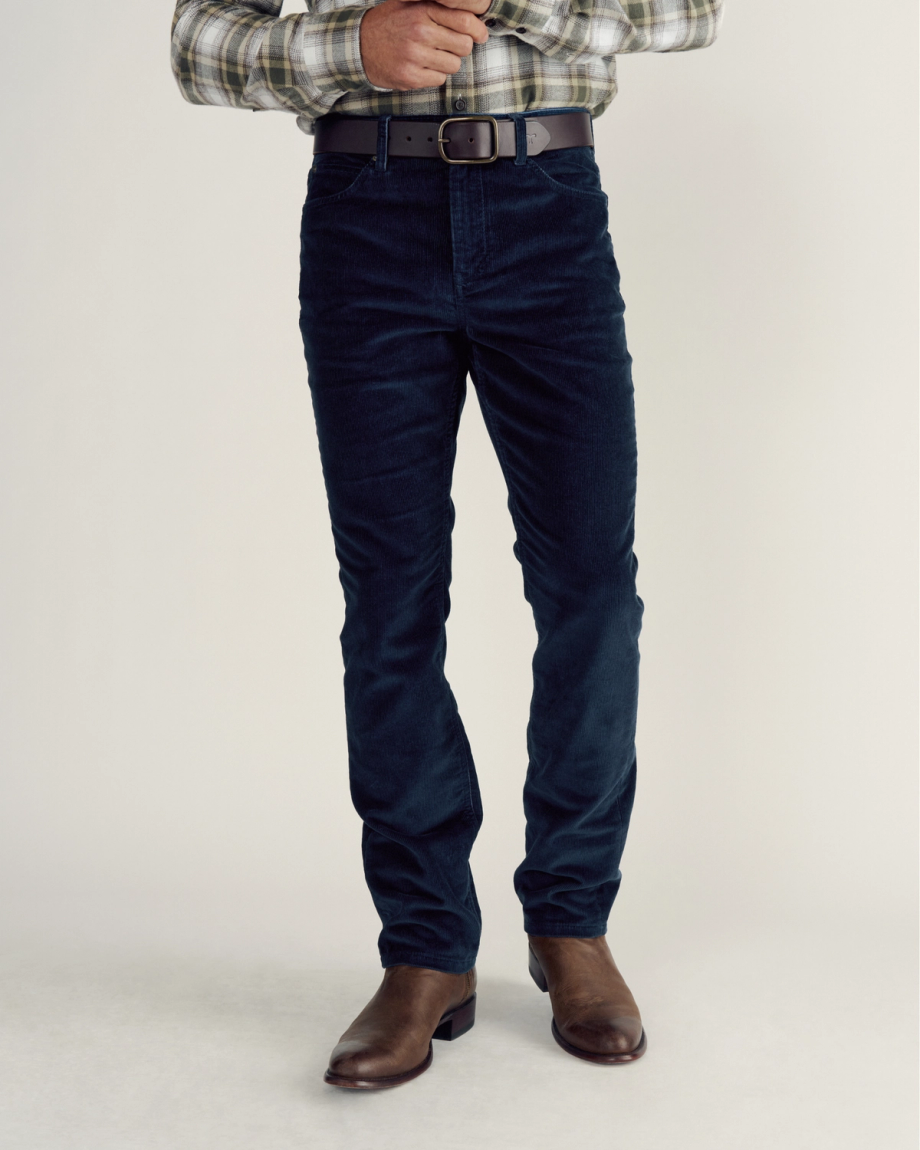 A person wearing a plaid shirt, dark blue corduroy pants, a belt, and brown leather boots stands against a plain background.