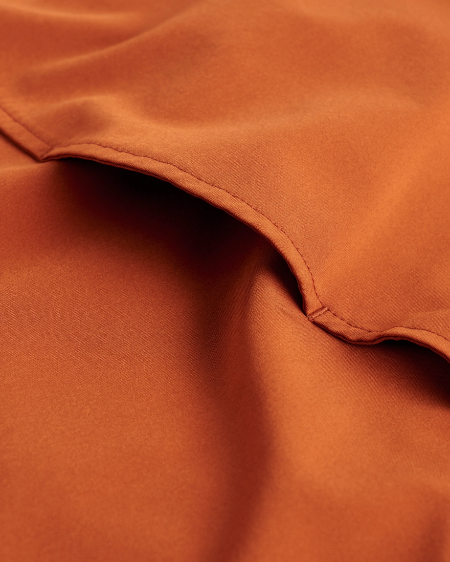 Close-up of rust-orange fabric with visible stitching and folds, highlighting the smooth texture and color of the material.